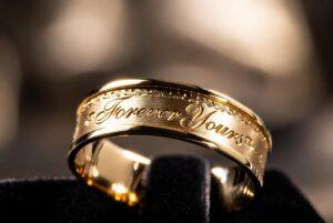 A luxurious gold ring engraving featuring the words "Forever Yours" in elegant script on a brushed metal band.